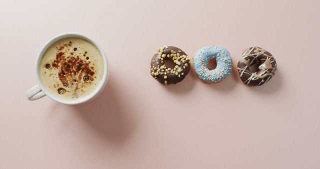 Flat lay showcasing a cappuccino next to three different donuts on a pink background. Perfect for use in food blogs, breakfast-themed marketing, and dessert advertisements. Highlights a casual and inviting coffee break setting.