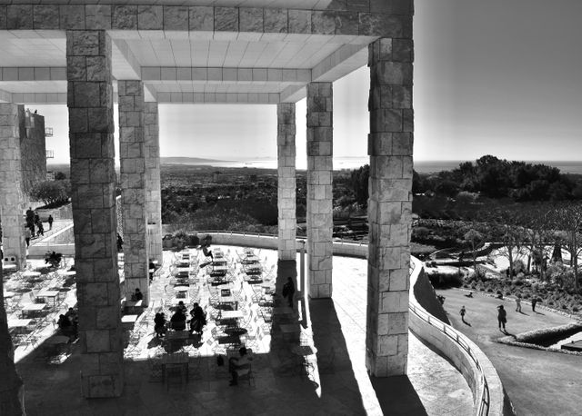 This photograph features a modern architectural structure with large stone pillars and an expansive outdoor seating area. The terrace offers a panoramic view of the surrounding landscape, encompassing both greenery and a distant cityscape. Several people are seated at picnic tables, enjoying the view, while others are walking along the pathways. This image is ideal for use in articles about architecture, urban design, outdoor recreation, or travel.