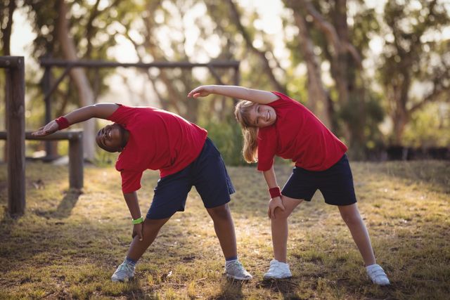 Two kids in red shirts and blue shorts are stretching during an outdoor boot camp. They are in a park-like setting, engaging in physical activity and promoting fitness and health. This image can be used for promoting children's fitness programs, outdoor activities, summer camps, and educational materials on physical education.
