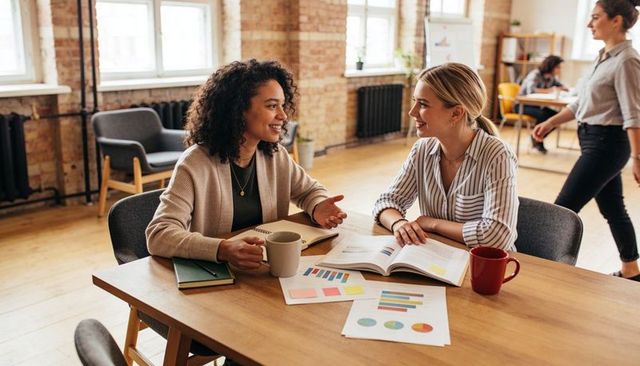 Two Women Collaborating Over Charts and Coffee at Modern Coworking Table, Reviewing Data