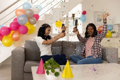 Diverse friends popping confetti and celebrating with balloons on living room sofa