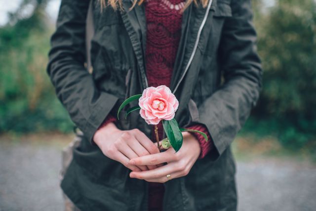 Woman stands outdoors holding a delicate pink flower. She wears an autumn-inspired green jacket and a sweater. Ideal for themes related to nature, relaxation, casual clothing, and outdoor activities. Perfect for using in advertisements, blogs, or social media posts highlighting autumn fashion or serene outdoor settings.