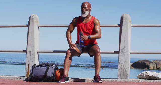 Senior man sitting on concrete railing near ocean after an outdoor workout. He is wearing a red tank top, black shorts, and holding a water bottle. His gym bag and a basketball are on the ground next to him. Ideal for use in materials promoting fitness, outdoor activities, health and wellness, and active lifestyles for seniors.