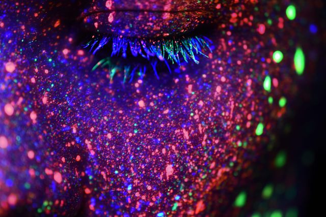 Close-up of person with face covered in colorful neon glow paint under UV light. The eye is closed, and the paint creates an abstract, vibrant look on the skin. This can be used for creative art projects, neon-themed party promotions, makeup design inspiration, and marketing materials for UV light products.