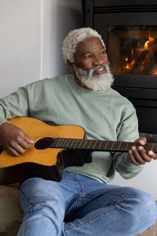 Senior african american man playing acoustic guitar by fireplace, cozy home music moment