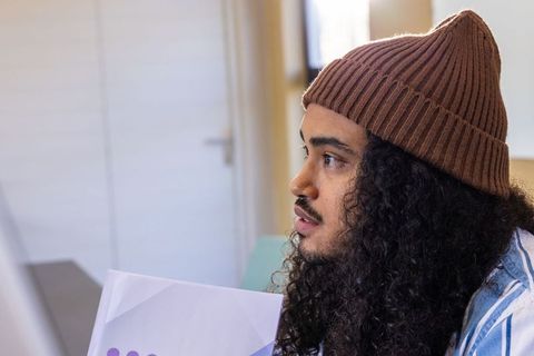 Young man wearing brown knit beanie holding purple folder listening attentively at desk