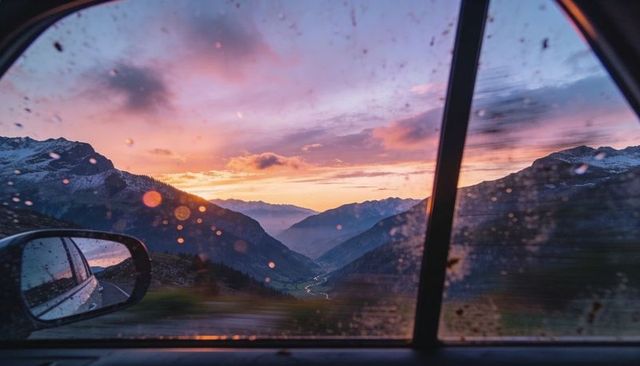 Watching alpine sunset through rain-dotted car window with side mirror road reflection