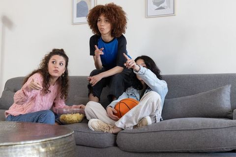 Teen friends watching game and pointing while sharing chips on gray couch with basketball