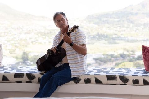 Middle-aged man playing acoustic guitar on window bench overlooking sunlit valley