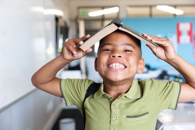 Portrait of smiling african american elementary boy holding book on head while standing in class. unaltered, education, playful, fun, happiness and school concept.