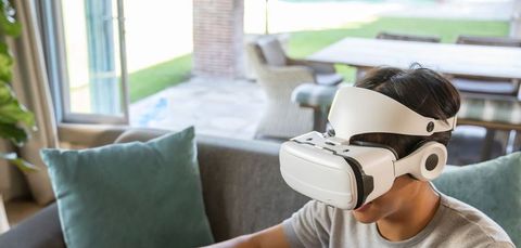 Asian man wearing white virtual reality headset sitting on gray sofa in sunlit living room