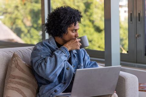 African american man sipping coffee while working remotely on laptop in cozy living room