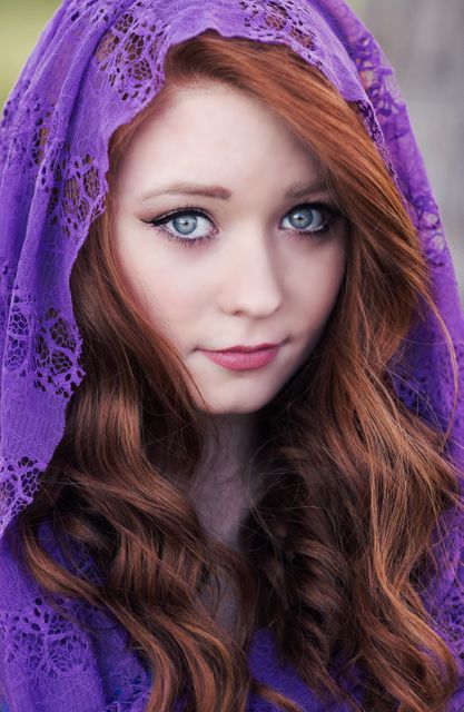 Young woman with striking red hair and piercing blue eyes wearing a detailed purple lace veil. Perfect for use in fashion editorials, beauty products advertisements, and websites focusing on style and lifestyle content. Also suitable for visual content related to themes of elegance, classic beauty, and artistic photography.