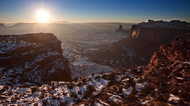 This stunning photograph captures the harmonious merge of warm sunset hues with the cool tones of a snow-covered canyon. The sun sets behind distant mountains, casting compelling shadows across the rugged terrain, highlighting the dramatic beauty of the rocky cliffs. Ideal for use in travel magazines, nature blogs, calendar designs, or inspirational posters showcasing natural landscapes and winter beauty.