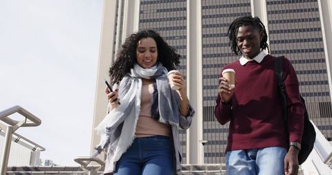 Young diverse friends walking down city stairs holding coffee cups and smartphone