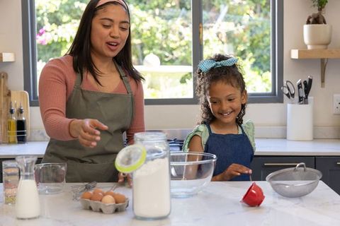 Asian mother teaching daughter baking in modern kitchen, mixing batter and smiling