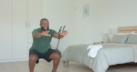 African american man doing squats in bedroom home workout with exercise bike nearby