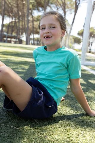 Young girl sitting on turf smiling near soccer goalpost in sunny park, relaxing casual