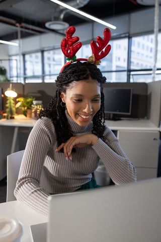 African-american woman wearing red reindeer antlers smiling and typing on laptop in office