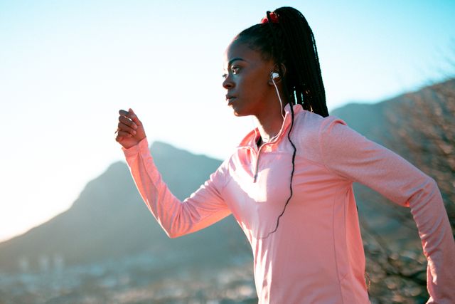 This image depicts a fit African American woman running outdoors while wearing wireless earphones. The background shows a scenic mountain view, highlighting the beauty of nature. Perfect for promoting fitness products, activewear, health and wellness blogs, motivational content and advertisements for outdoor activities.