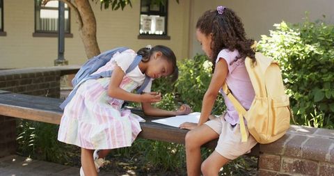 Primary school girls collaborating on homework outdoors on wooden bench with backpacks