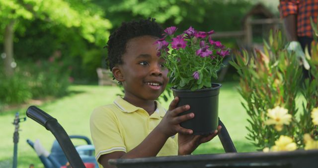 Young African American boy happily holding a purple flower pot while in a garden. Suitable for themes of gardening, childhood activities, outdoors, nature, and family fun. Ideal for educational materials, lifestyle blogs, and advertisements promoting family-friendly activities.