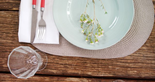 A neatly arranged table setting features a plate with a delicate floral decoration, silverware wrapped in a white napkin, and an overturned wine glass, with copy space. The rustic wooden surface adds a warm, natural touch to the elegant dining presentation.