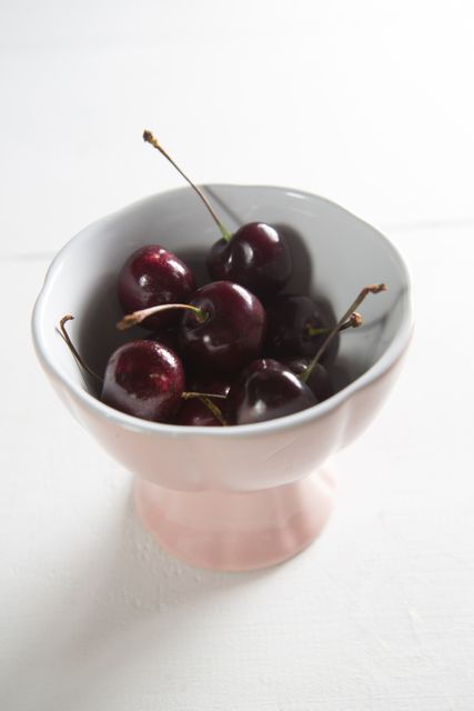 Fresh cherries in a pink bowl on a white table. Ideal for use in food blogs, healthy eating articles, summer recipes, and advertisements for organic produce. The vibrant colors and simple composition make it suitable for kitchen decor and culinary magazines.