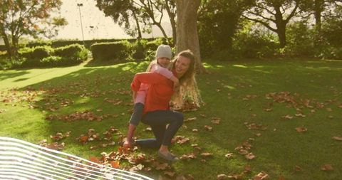 Mother giving daughter piggyback in autumn park with sunlight and fallen leaves