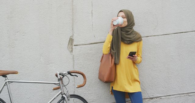 Young woman wearing hijab standing against wall, holding a coffee cup and smartphone, with bicycle nearby. Ideal for illustrating modern lifestyles, technology use, city living, or cultural diversity themes.