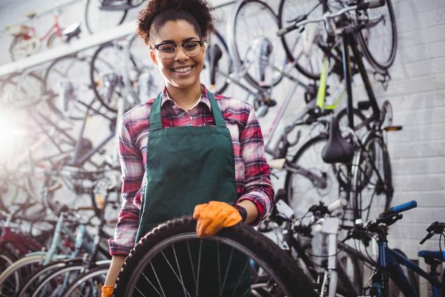 Female mechanic smiling while holding a bicycle tire in a workshop filled with bikes. Ideal for use in content related to bicycle repair services, professional mechanics, women in trades, and small business promotions.
