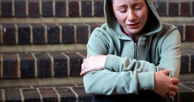 Teenager in a green hoodie, appearing sad and reflective, sitting alone on brick stairs. This image can be used in articles and campaigns related to mental health awareness, adolescent challenges, support for youth, loneliness, and emotional wellbeing.