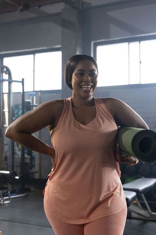 African american woman smiling carrying rolled yoga mat in gym wearing pink activewear