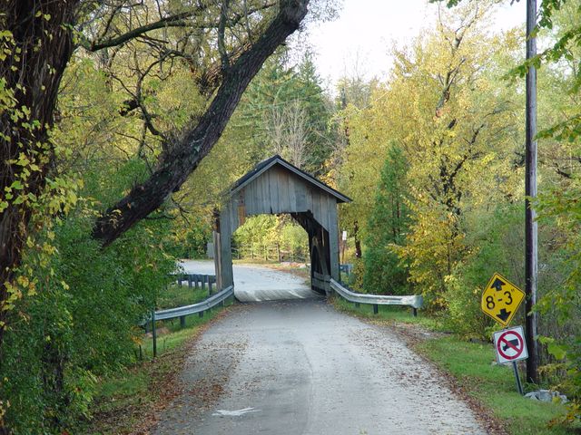 Idyllic rural scene featuring a rustic covered bridge on a tranquil forest road during autumn. Ideal for illustrating peaceful countryside travel, historical landmarks, scenic drives, or seasonal changes. Perfect for tourism ads, brochures, and websites aiming to capture the charm of rural destinations.