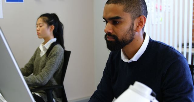Two focused colleagues are working independently at their desks in an office environment. A bearded man in a dark sweater is looking at his computer monitor, while an Asian woman, in a gray sweater, is concentrating on her screen in the background. Ideal for depicting modern office work, productivity, and professional settings.