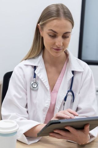 Female clinician reviewing patient records on tablet wearing lab coat and stethoscope