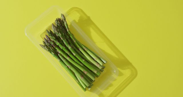 This image depicts a bunch of fresh green asparagus stalks neatly lined in a clear plastic container against a vibrant yellow background. The vivid colors emphasize the freshness and healthiness of the vegetables. This can be perfect for use in nutrition and healthy living magazines, diet blogs, food marketing campaigns, and any content that promotes a healthy lifestyle. The striking yellow background adds a lively and modern touch to culinary presentations or restaurant menus.