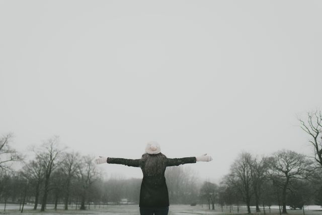 Person standing alone outdoors with arms outstretched in wintertime, enjoying freedom and serenity. Bare trees and overcast sky create peaceful, reflective atmosphere. Suitable for themes like mindfulness, relaxation, outdoor activity, and seasonal content.