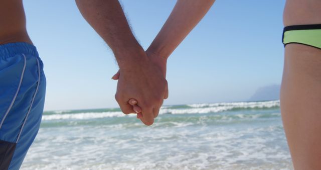 A Caucasian couple holds hands on a sunny beach, with copy space. Their intimate gesture and the serene ocean backdrop evoke a sense of romantic connection and leisure.