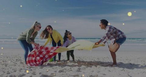 Friends holding bright blankets on sunlit beach preparing picnic and laughing