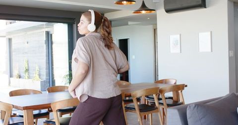 Woman standing in modern dining area listening to music with white over-ear headphones