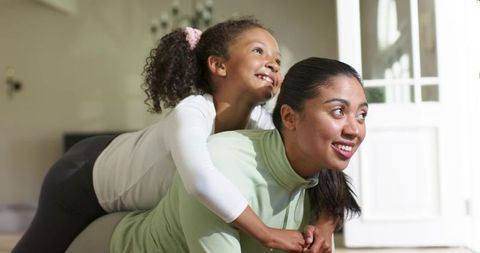 Mother carrying daughter on back while smiling and playing indoors in bright cozy home