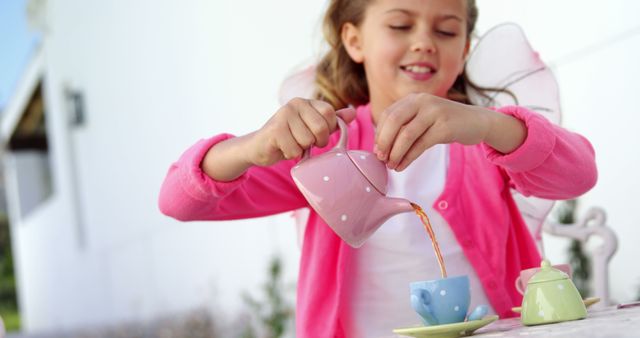 A young Caucasian girl is playfully pouring tea from a pink teapot into a small cup during a pretend tea party, with copy space. Her imaginative playtime captures a moment of childhood innocence and joy.