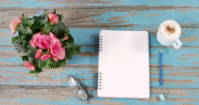A vibrant bouquet of pink roses sits next to an open notebook with a pencil, glasses, and a cup of coffee on a rustic blue wooden table, with copy space. It's an inviting scene for writing, planning, or enjoying a quiet moment.