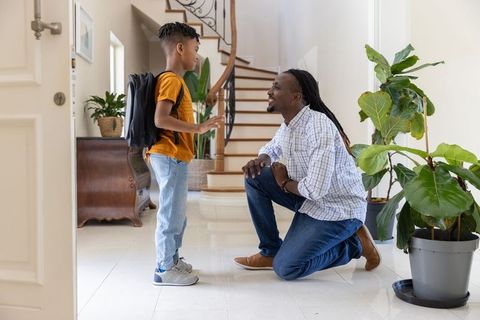 Father kneeling and talking to son with backpack near curved staircase in home entryway