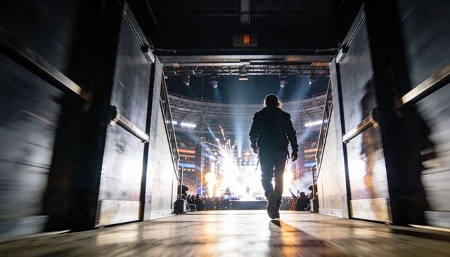 Silhouette performer walking backstage toward pyrotechnic stage lights and roaring crowd