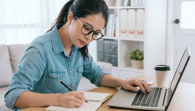 Focused remote worker taking notes while using laptop in bright tidy home workspace