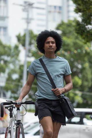 African american man walking city street with road bike, wearing teal tee and crossbody bag