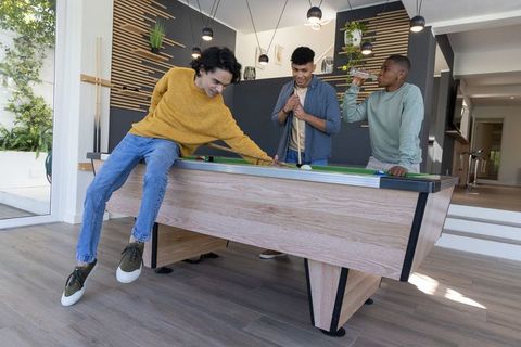 Diverse friends playing pool in modern living room with wooden billiards table