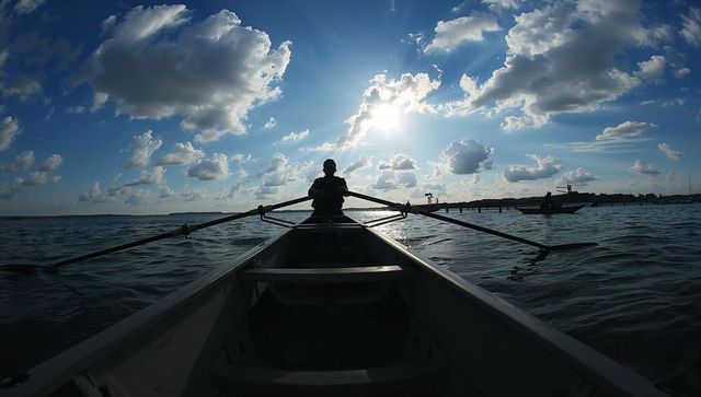 Silhouetted rower paddling single scull on sunlit bay under dramatic cloud sky